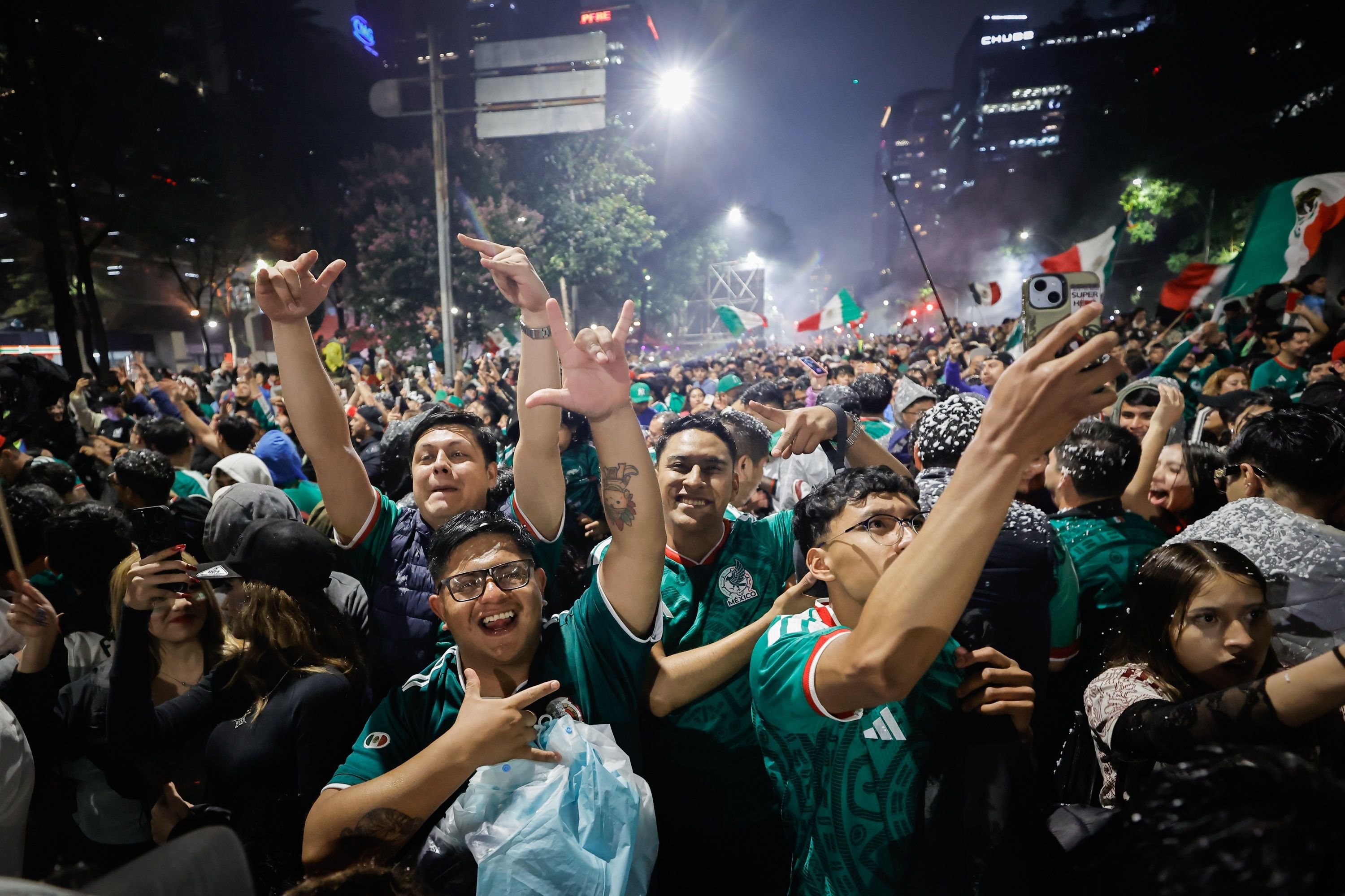 A large nighttime crowd wearing green Mexico jerseys cheers and poses for a selfie, with Mexican flags waving and city buildings visible behind them.