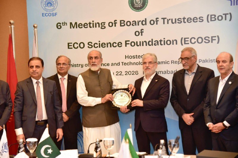Seven men in formal attire pose on stage as two exchange a plaque at the 6th Meeting of the Board of Trustees of the ECO Science Foundation, with Pakistani flags visible.