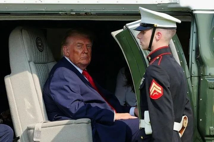 A man in a dark suit and red tie sits inside a helicopter, facing a uniformed Marine wearing a white cap and dress blues with red chevrons standing in the open doorway.