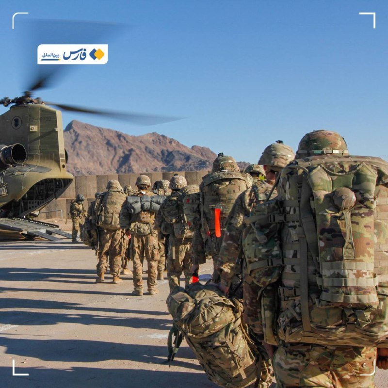 Soldiers in camouflage uniforms and helmets walk in a line carrying large rucksacks toward a green military helicopter with spinning rotors on a dusty terrain with mountains in the background.