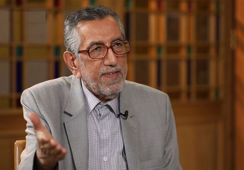 A gray-haired, bearded man wearing glasses, a gray blazer, and a checkered shirt gestures with his hand during an interview.