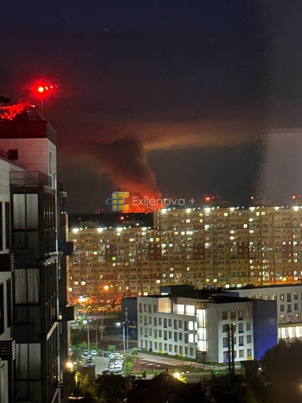 A nighttime cityscape shows lit apartment buildings and a lower commercial structure, with an orange glow and dark smoke rising in the distance.