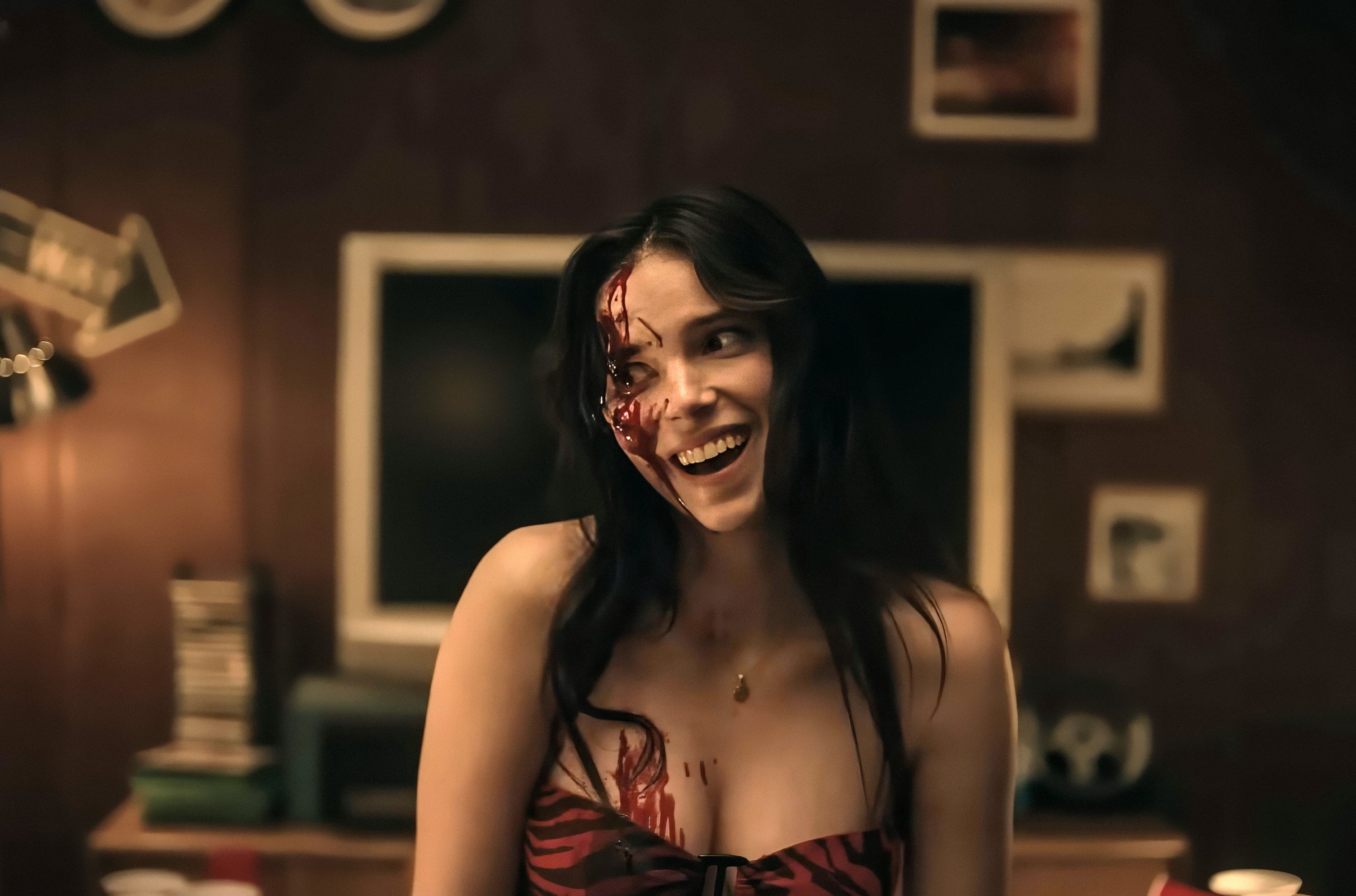 A woman with long dark hair and a blood-smeared face smiles widely in a dimly lit room with framed pictures on the wall.