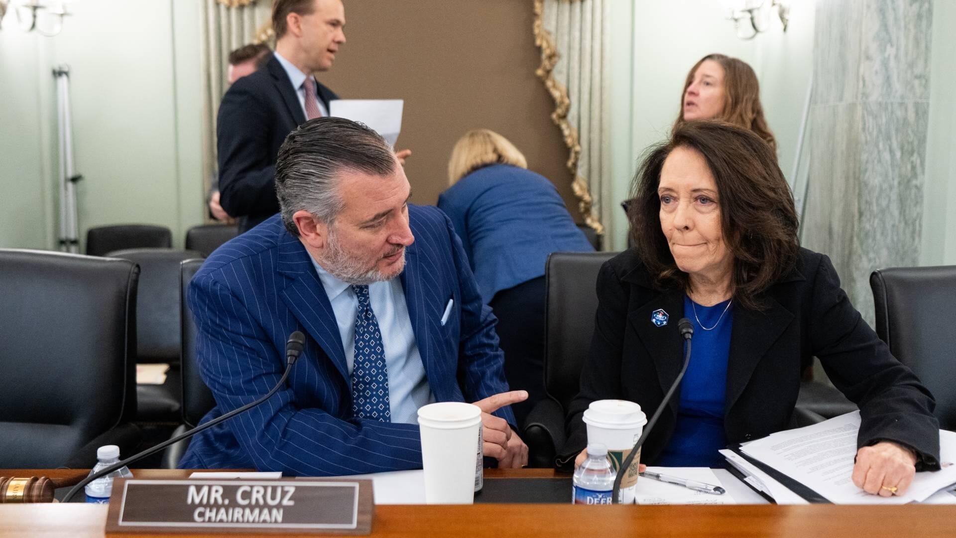 A man in a blue pinstripe suit sits at a wooden dais with a "MR. CRUZ CHAIRMAN" nameplate, conferring with a woman in a black blazer beside coffee cups and documents.