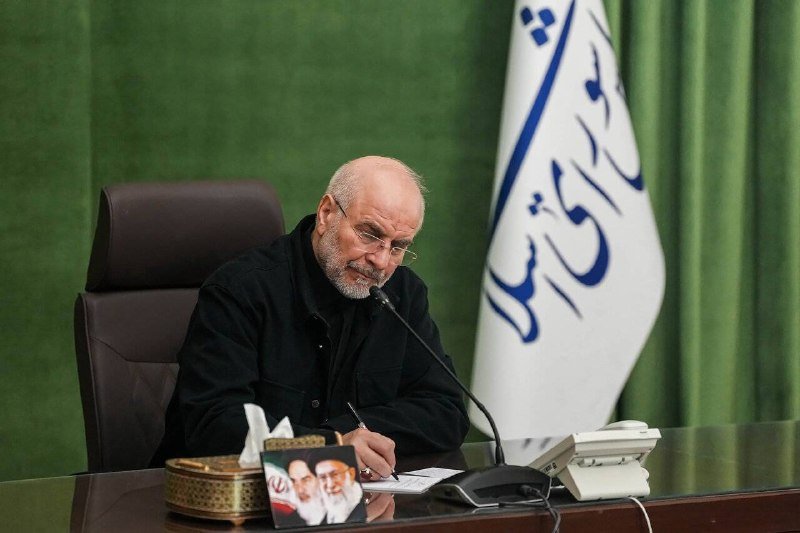 A bespectacled, bearded man in black sits at a desk writing with a pen, with a Persian-script flag and framed photos visible nearby.