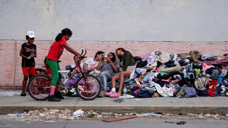 Children rest beside a large pile of discarded clothing on a sidewalk while a girl with a red bicycle and a boy in a white cap stand nearby.
