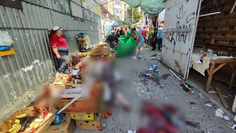 A crowded outdoor market alley shows vendors selling produce from wooden crates and boxes, with shoppers walking between stalls beneath a shaded canopy.