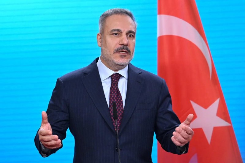 A gray-haired man in glasses and a suit speaks at a microphone, with Arabic text and a logo overlaid on the image.