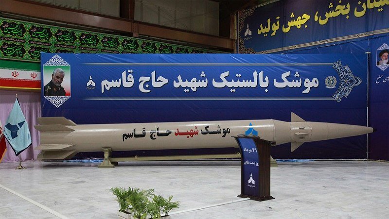A large missile labeled with Persian script and a portrait is displayed indoors beneath a blue banner with Persian text, flanked by Iranian flags.