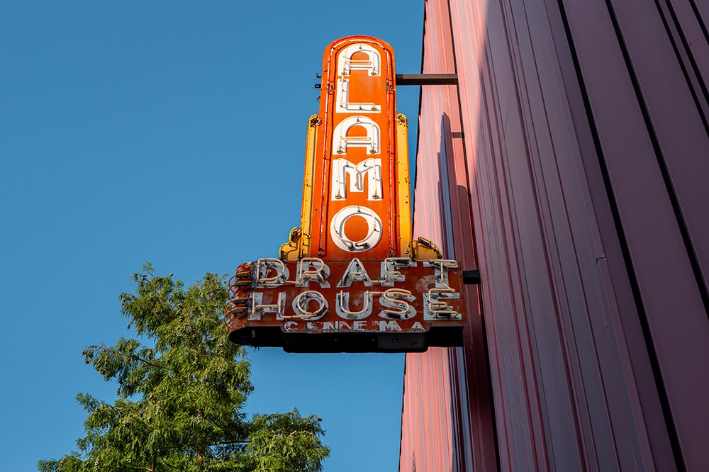 A weathered, vintage orange-and-yellow neon sign reading "ALAMO DRAFTHOUSE CINEMA" extends from a building's exterior against a clear blue sky.