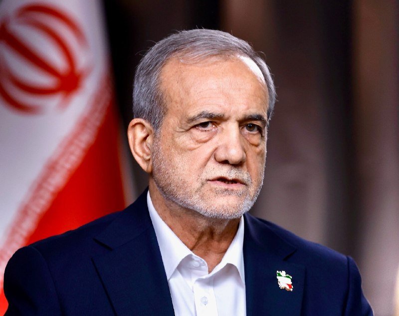 An older man with gray hair and a beard, wearing a dark suit and white shirt with an Iranian flag pin, speaks in front of an Iranian flag.