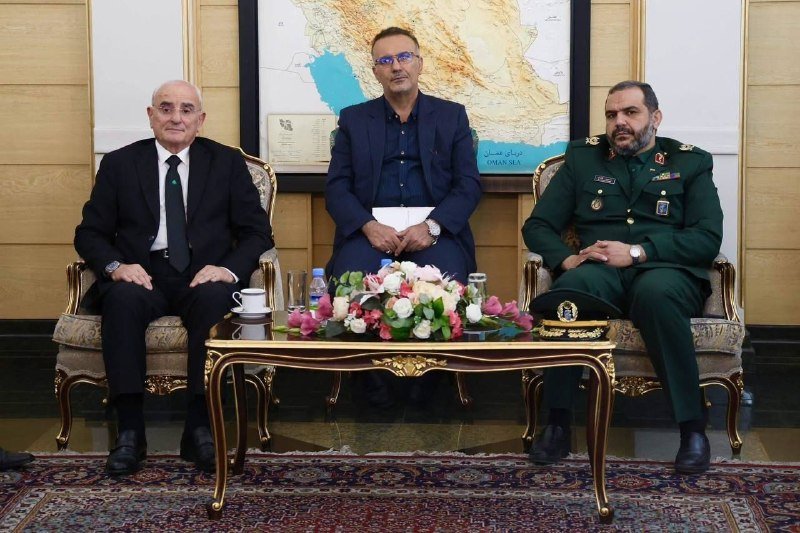Three men sit formally posed behind a coffee table with a floral arrangement, in front of an Oman Sea regional map.
