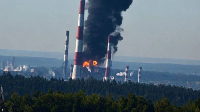 Thick black smoke and flames billow from an industrial facility with tall red-and-white striped smokestacks, set against a forested landscape.