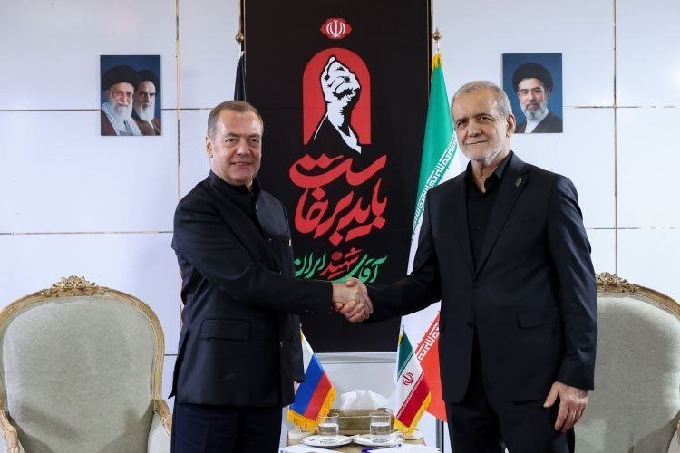 Two men in dark suits shake hands in front of Iranian, Russian, and Armenian flags, framed by portraits of clerics and a banner with Persian script and a raised fist graphic.