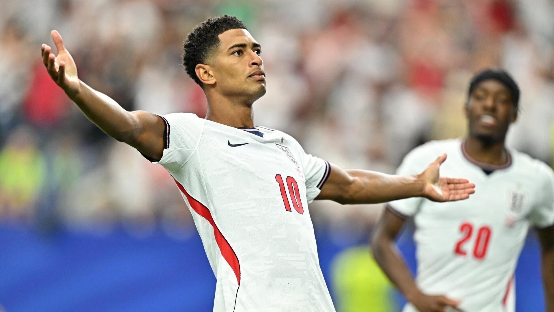 A soccer player wearing a white jersey with the number 10 and a team crest stands with arms outstretched in celebration, while a teammate wearing number 20 approaches from behind.