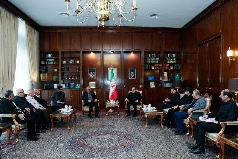 Two seated officials face each other in a wood-paneled meeting room flanked by an Iranian flag, portraits, and bookshelves, with attendees seated along both walls.