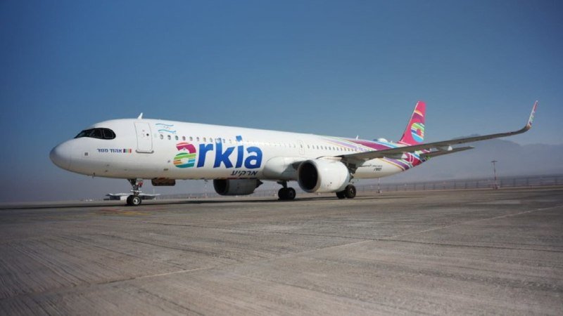 A white Arkia passenger jet with colorful tail livery sits on an airport tarmac under a clear blue sky.