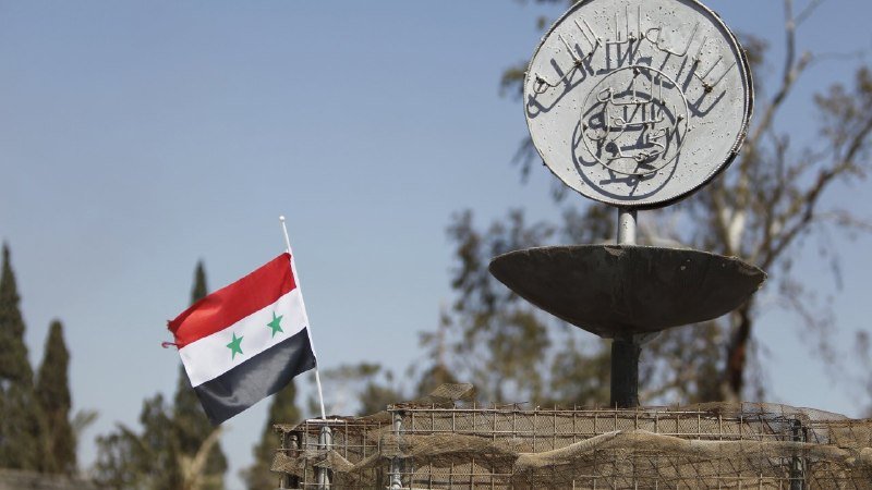 A Syrian flag flies beside a sandbagged structure topped with a circular sign displaying Arabic calligraphy, set against a clear blue sky with trees in the background.