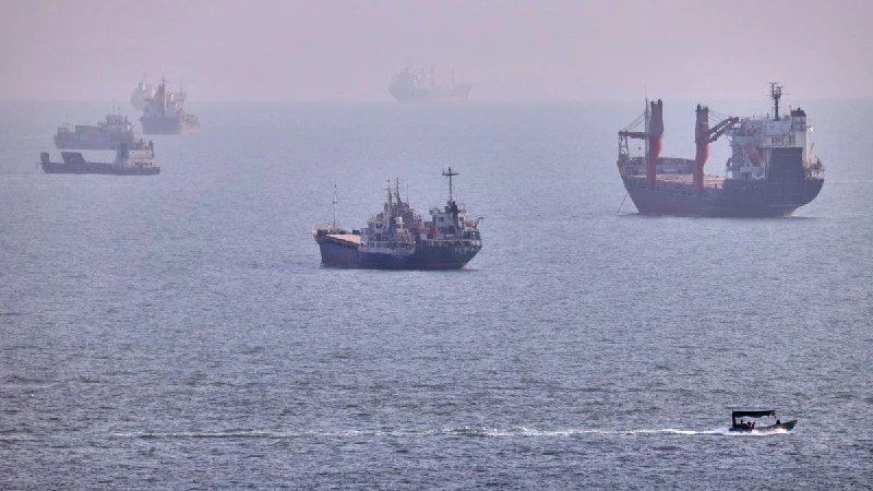 Several cargo and industrial ships sit anchored in hazy waters while a small motorboat cruises past in the foreground.
