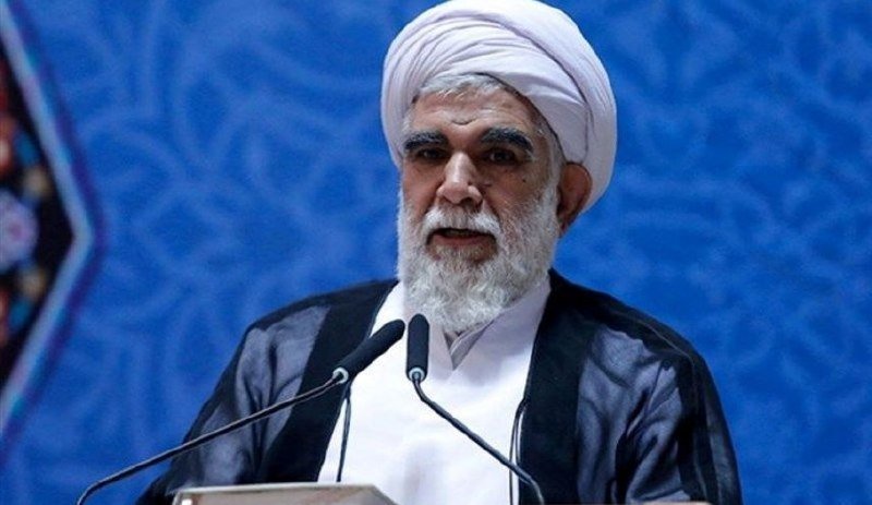 A bearded man wearing a white turban and dark robe speaks at a podium with microphones, set against a blue patterned backdrop.