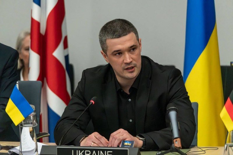 A man in a black suit speaks into a microphone at a conference table with a "UKRAINE" placard, flanked by Ukrainian, British, and German flags.