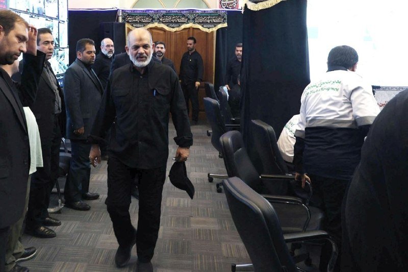A white-bearded man in clerical attire speaks at a podium with microphones, flanked by posters bearing Arabic script, Iranian and Lebanese flags, and an image of the Dome of the Rock.