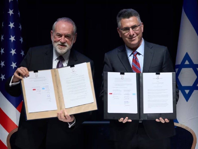 Two men in suits hold open folders displaying signed documents, standing before the U.S. and Israeli flags.