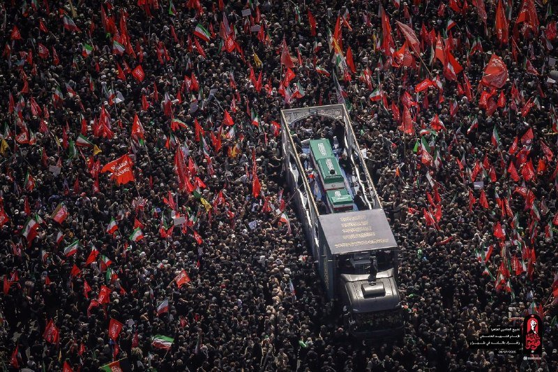 An aerial view shows a massive crowd waving red flags and Iranian flags surrounding a truck carrying green-draped coffins marked with Arabic script.
