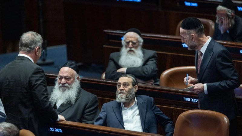 Several men in suits, some wearing kippahs, converse in a wood-paneled legislative chamber with Hebrew text visible on the desks.