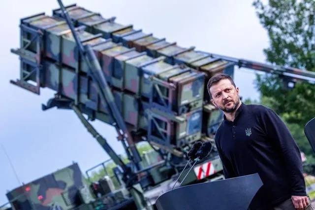 A bearded man wearing a dark shirt with a small crest stands at a podium beside a camouflage-painted military vehicle equipped with an elevated launcher of boxy missiles, with trees in the background.