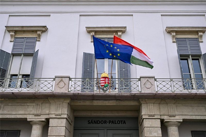 The European Union and Hungarian flags fly above a balcony displaying the Hungarian coat of arms at the entrance of a building marked "SÁNDOR-PALOTA."