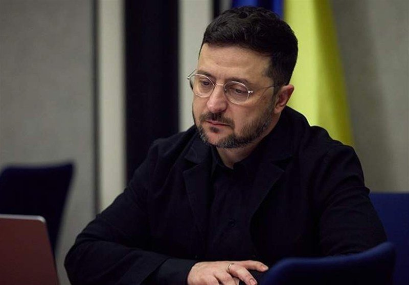 A bearded man wearing glasses and a dark suit sits looking downward beside a laptop, with blue and yellow flags visible in the background.