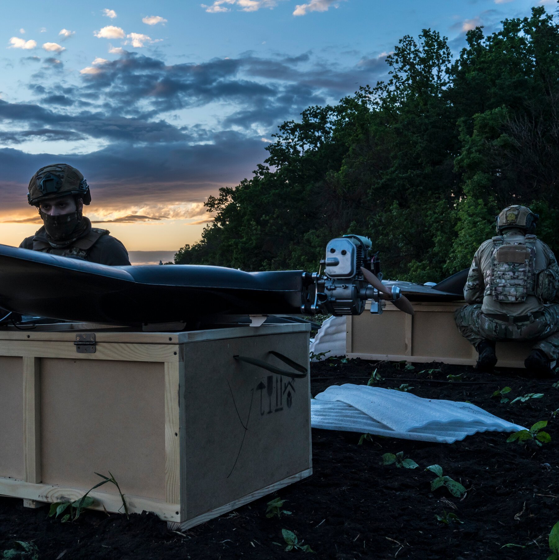 Two masked soldiers in tactical gear tend to a large drone resting atop wooden crates in a field at dusk.