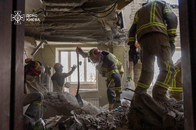 Rescue workers in protective gear clear rubble and debris from inside a heavily damaged building, with a logo reading "ДСНС КИЄВА" visible in the corner.
