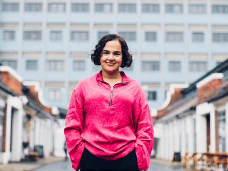 A woman with short dark curly hair, wearing a bright pink half-zip fleece, stands smiling at the camera in front of a modern building.