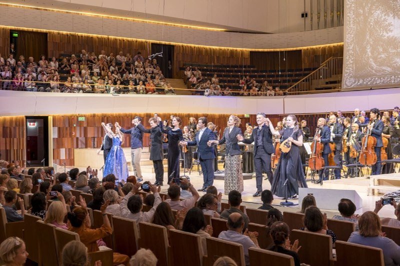 Performers and musicians stand on a brightly lit concert hall stage, taking bows before a packed audience holding up phones to capture the moment.