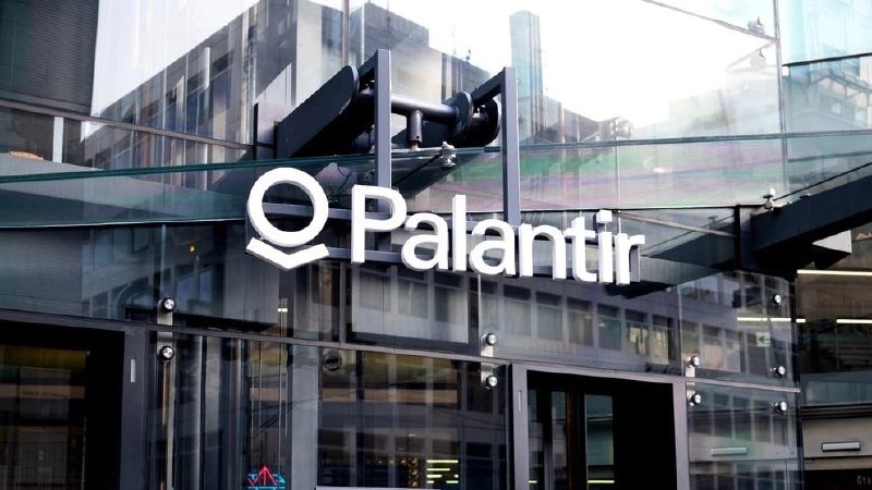 A "Palantir" sign with a logo is mounted above glass entrance doors of a modern office building.