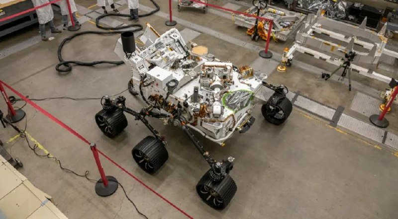 A six-wheeled robotic rover prototype with exposed wiring and components sits on a concrete floor, cordoned off by red stanchions and ropes in an industrial facility.