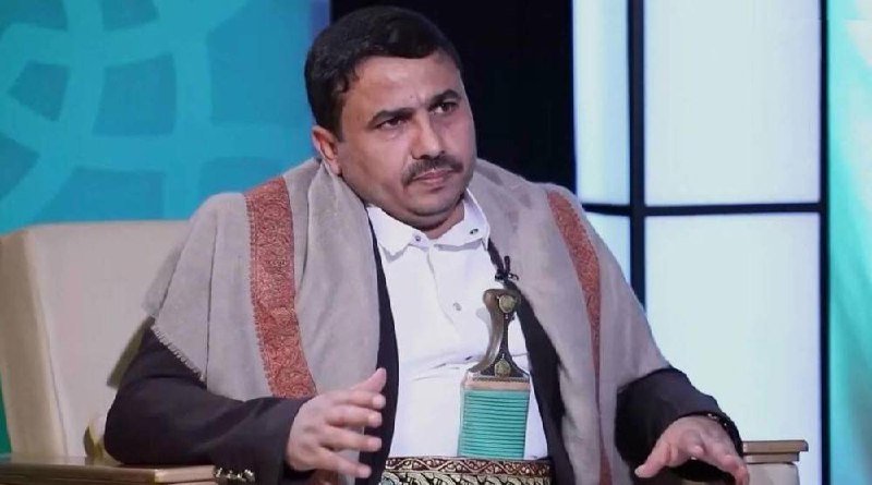 A man wearing a white shirt and a shawl sits in a studio chair, gesturing with his hand while appearing to speak during an interview on a teal-colored set.