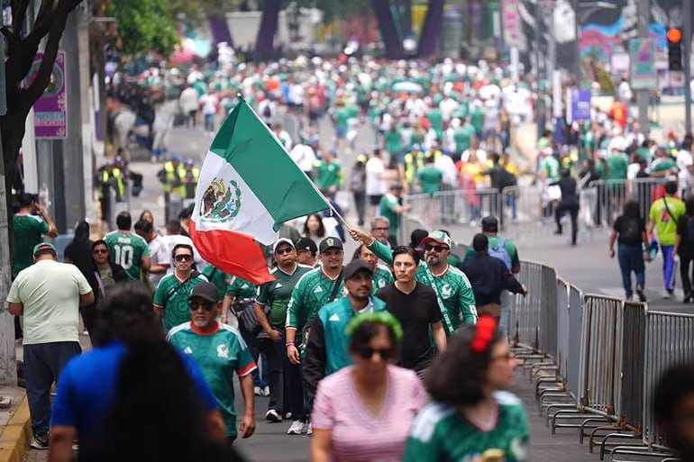 A crowd of people, many wearing green jerseys, walks down a street behind metal barricades, with one person prominently waving a large Mexican flag.