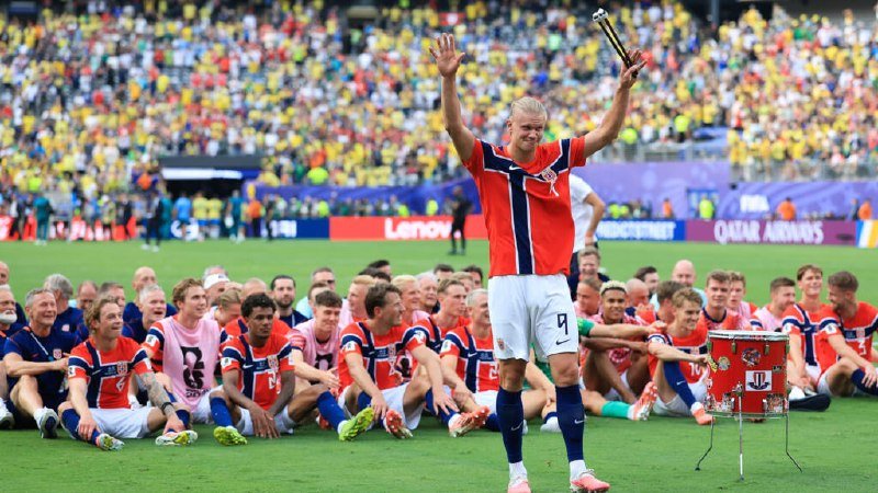 Blonde soccer player in a red, white, and blue jersey (#9) raises a trophy overhead while seated teammates cheer on a stadium field.