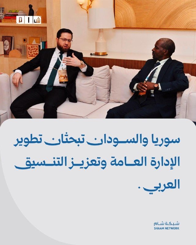 Two suited men wearing conference badges sit on a cream sofa in a modern lounge, gesturing while conversing, above Arabic text and a Shaam Network logo.