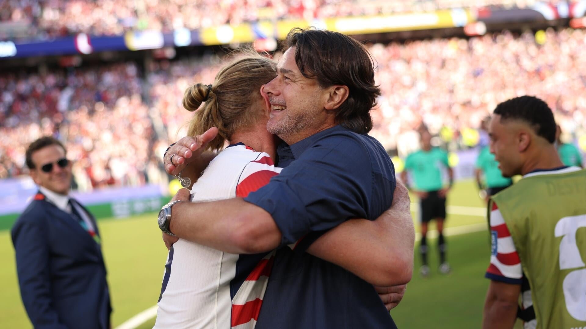 U.S. men's soccer players in red-and-white striped jerseys and substitute bibs celebrate together on the field in front of a packed stadium crowd.