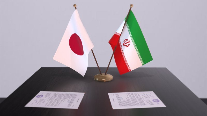 Two small desk flags—the white-and-red Japan flag and the green, white, and red Iran flag—stand crossed on a gold base atop a dark table, with two printed documents placed in front of them.