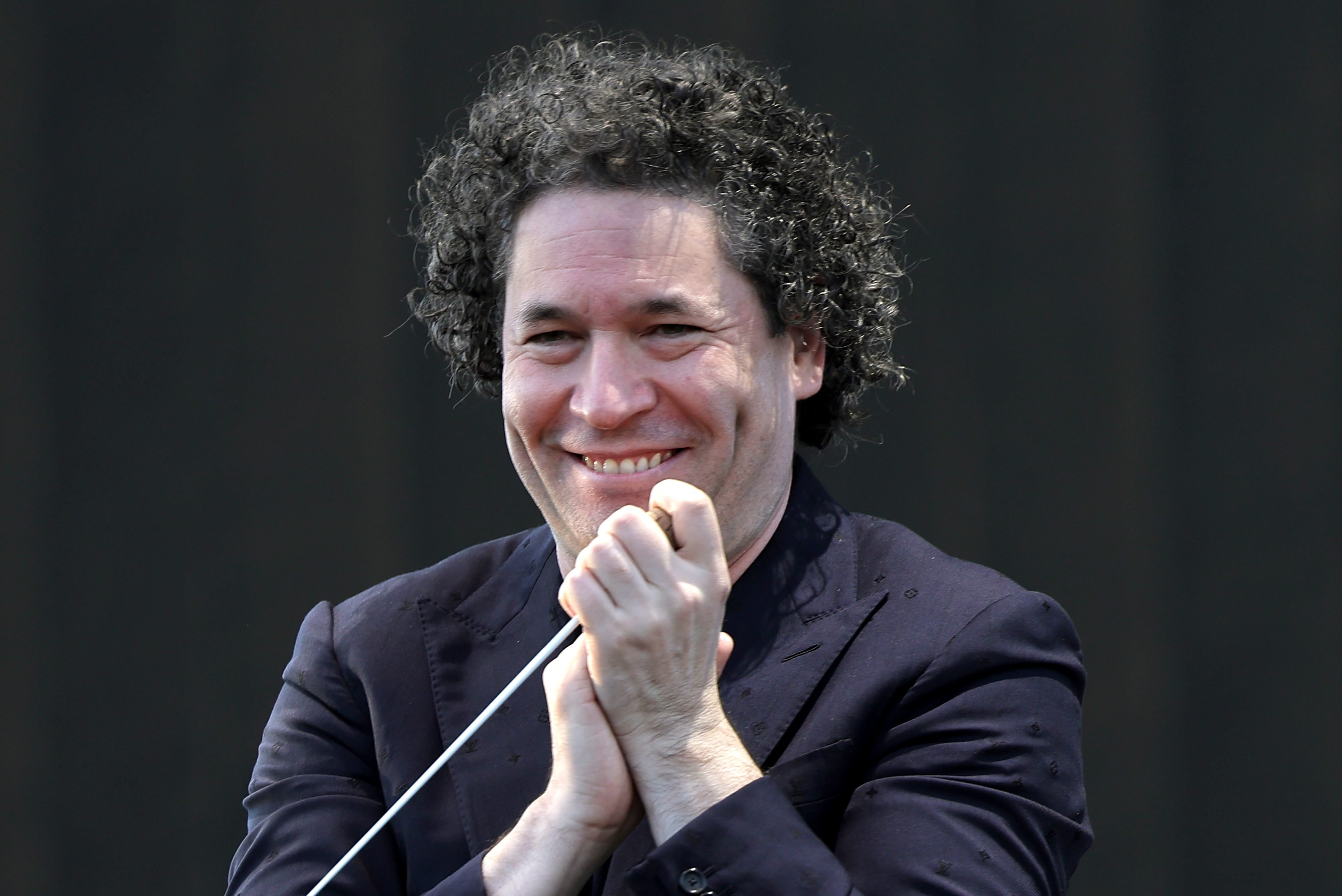 A curly-haired conductor in a dark patterned blazer smiles broadly while gripping a baton against a black background.
