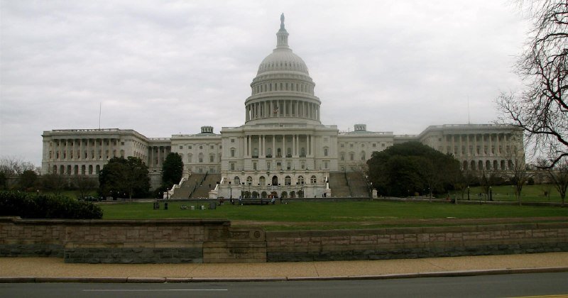 A large white neoclassical stone building with columns, an American flag on top, and a sculpted eagle above the entrance, surrounded by trees and a black bollard-lined walkway.