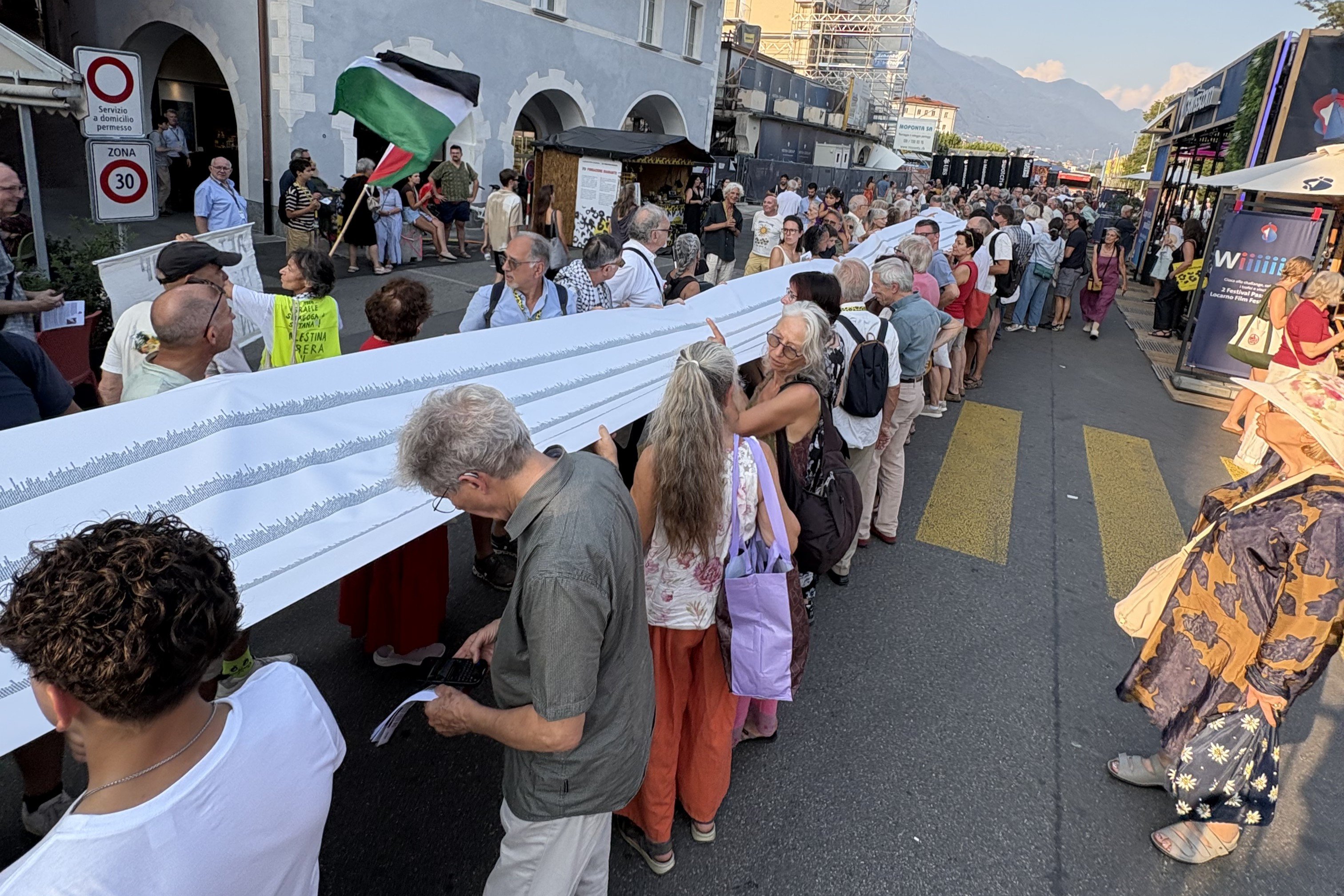 A crowd lines a street holding a long printed banner, while a person waves a Palestinian flag nearby at what appears to be an outdoor festival.