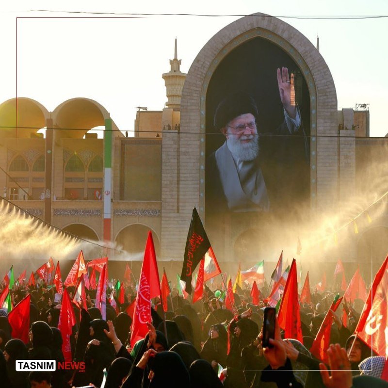 A large crowd waves red and Iranian flags before a massive archway portrait of a bearded cleric in a black turban, with a domed building behind.