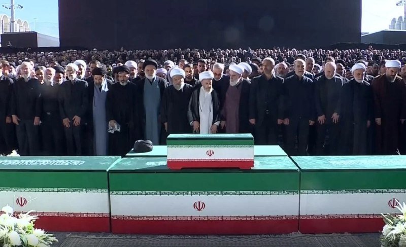 Clerics in white and black turbans and robes stand solemnly behind coffins draped in the Iranian flag, addressing a large gathered crowd at an outdoor ceremony.