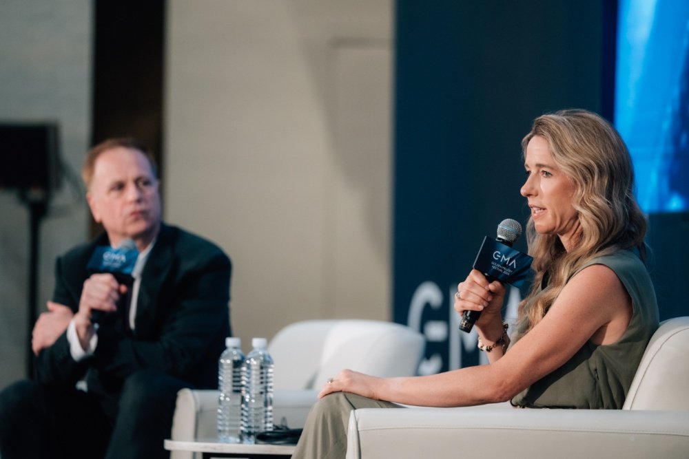 A blonde woman in an olive dress speaks into a microphone while a man in a dark suit listens, seated at a stage panel with water bottles.